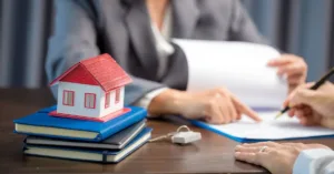 Pessoa assinando contrato com maquete de casa sobre livros, representando a documentação para compra e venda de imóveis.