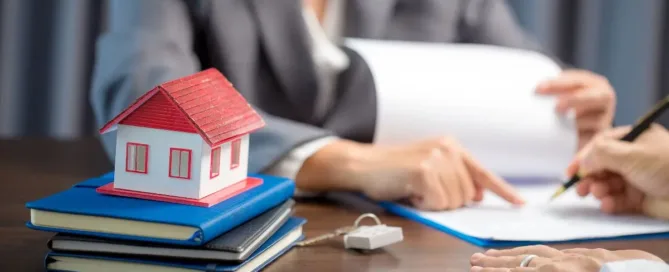 Pessoa assinando contrato com maquete de casa sobre livros, representando a documentação para compra e venda de imóveis.