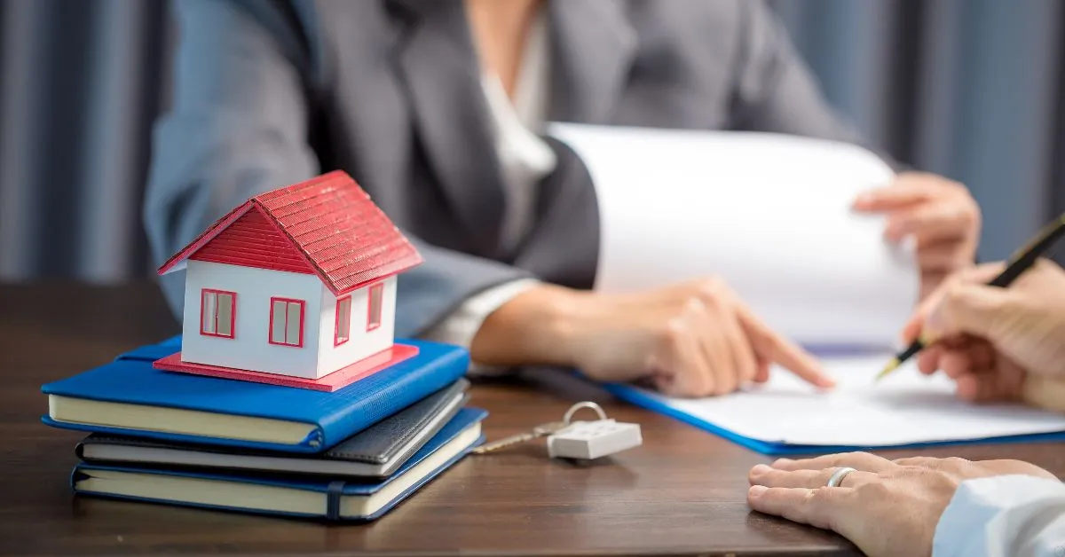 Pessoa assinando contrato com maquete de casa sobre livros, representando a documentação para compra e venda de imóveis.