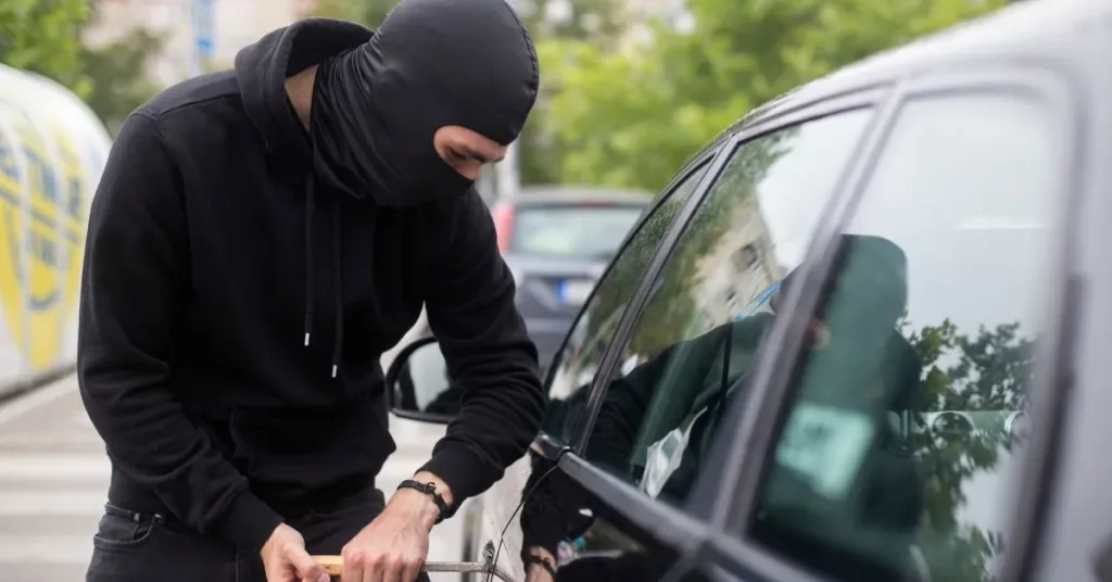 Homem mascarado tentando arrombar um carro estacionado durante o dia, representando furto em estacionamento de loja.
