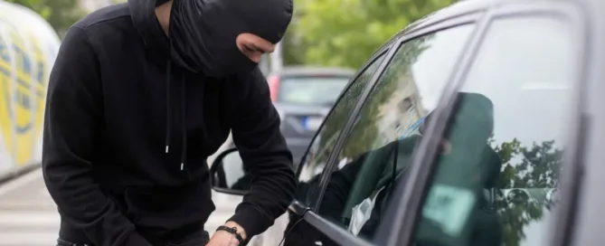Homem mascarado tentando arrombar um carro estacionado durante o dia, representando furto em estacionamento de loja.