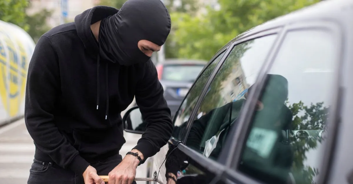 Homem mascarado tentando arrombar um carro estacionado durante o dia, representando furto em estacionamento de loja.