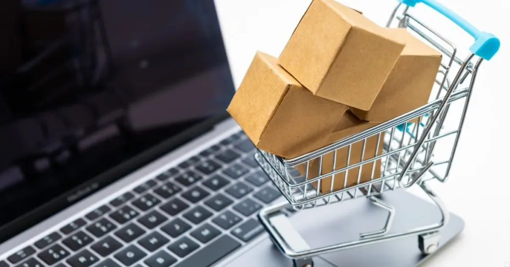 Carrinho de compras com caixas sobre teclado de notebook, representando compras online e troca de produto com defeito.