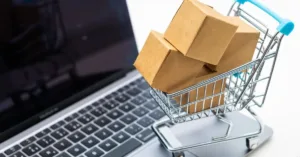Carrinho de compras com caixas sobre teclado de notebook, representando compras online e troca de produto com defeito.