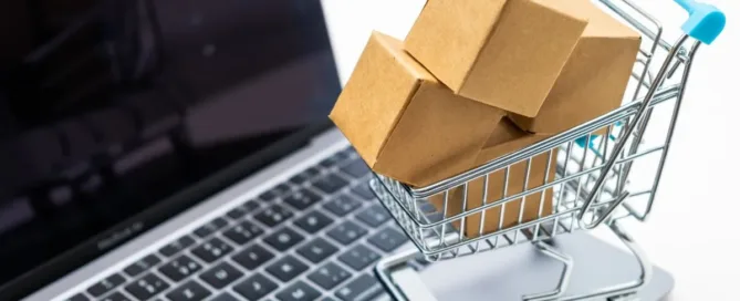 Carrinho de compras com caixas sobre teclado de notebook, representando compras online e troca de produto com defeito.