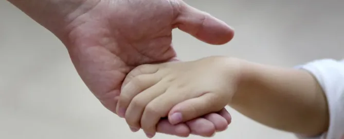 Mão de adulto segurando a mão de uma criança, simbolizando vínculo afetivo e proteção parental