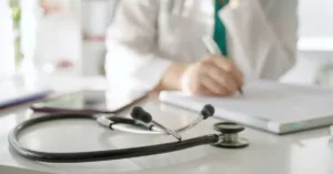 Estetoscópio sobre mesa médica com profissional ao fundo preenchendo prontuário psiquiátrico