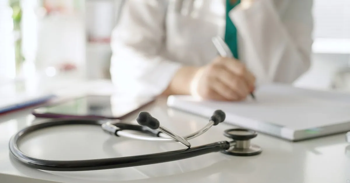 Estetoscópio sobre mesa médica com profissional ao fundo preenchendo prontuário psiquiátrico
