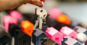 Chave de imóvel sendo entregue sobre maquete de casas coloridas, representando locação por temporada.