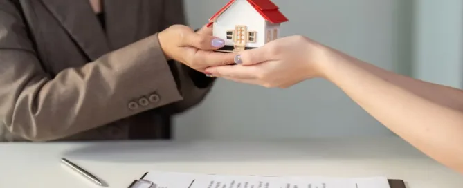 Pessoa entregando uma miniatura de casa em negociação imobiliária por cessão de direitos, com contrato sobre a mesa.