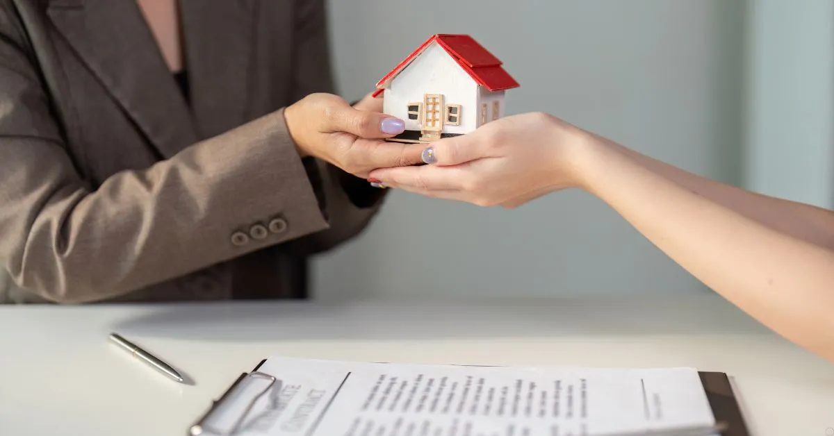 Pessoa entregando uma miniatura de casa em negociação imobiliária por cessão de direitos, com contrato sobre a mesa.